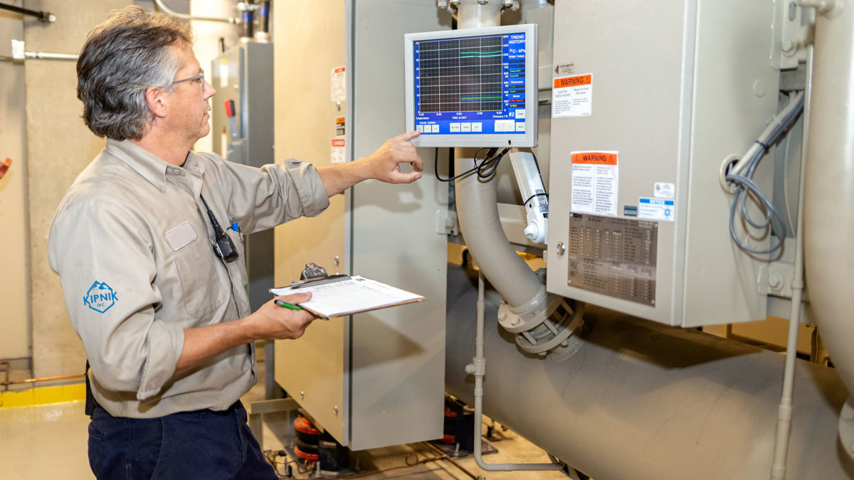 HVAC Technician inside a boilder room pushes a button on a digital monitoring system while holding a clipboard.